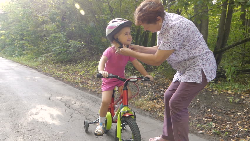 Женщина застегивает детский шлем на девочке в розовом, которая сидит на велосипеде