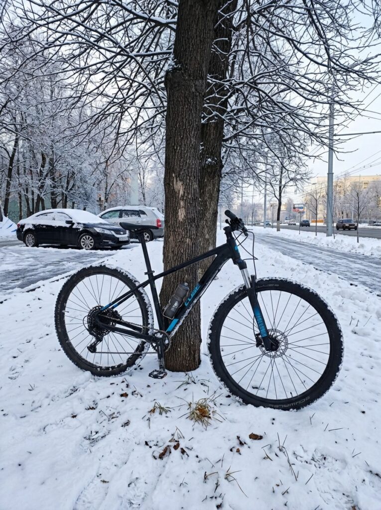 Передний дисковый тормоз и вилка велосипеда зимой Хардтейл Cube с вилкой RockShox и передним дисковым тормозом у дерева на снегу, рядом заснеженные машины